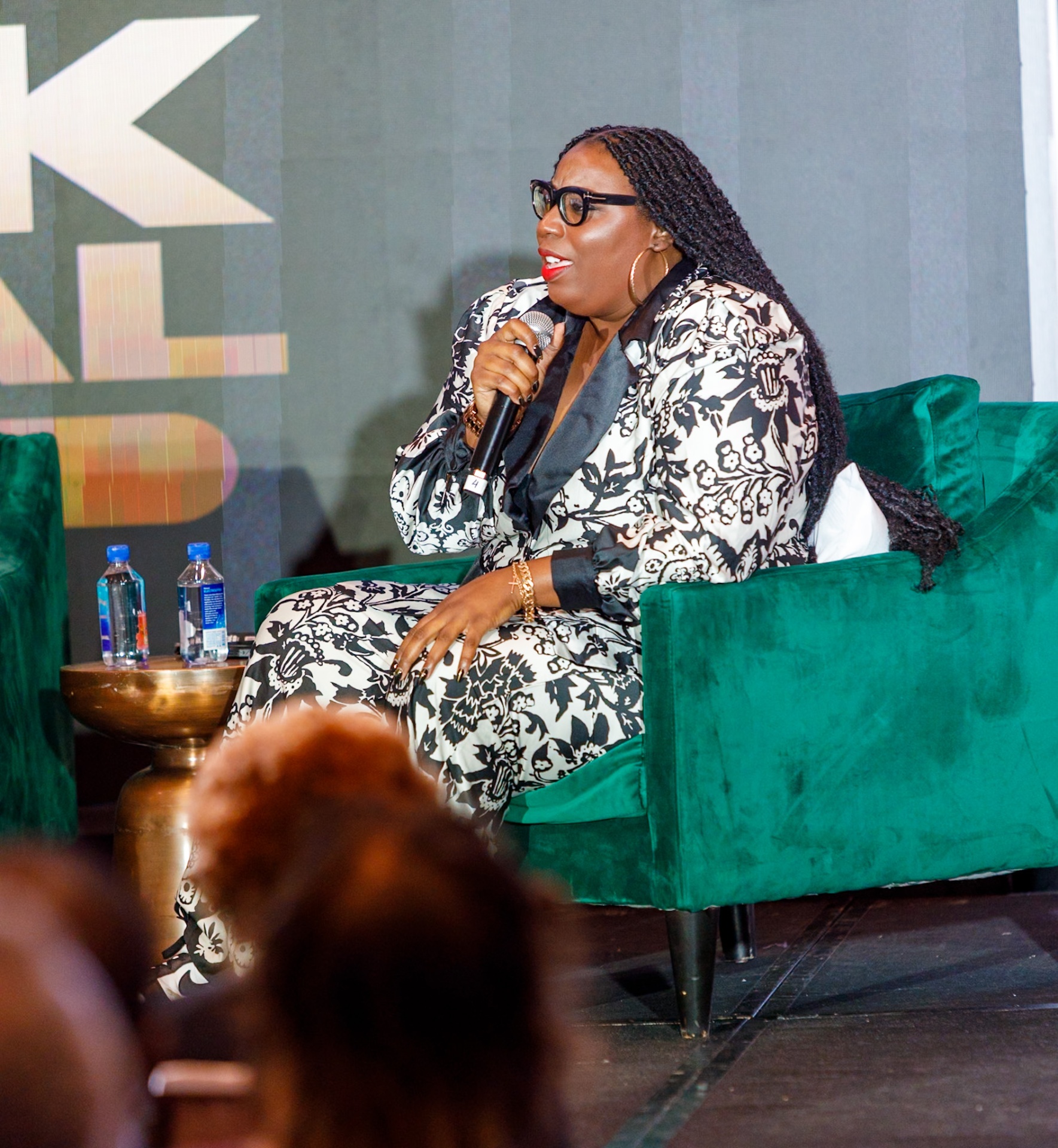Madam Prezident on stage in emerald velvet armchair, mic in hand, commanding the room